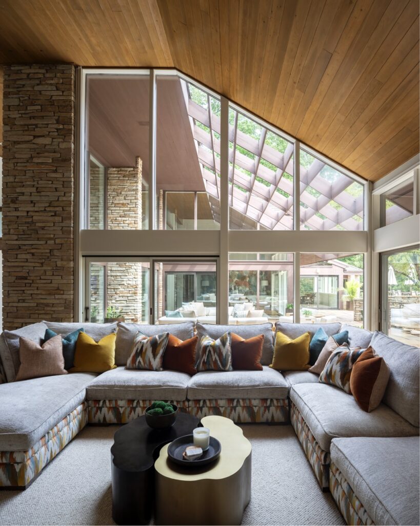 A large sectional couch hugs the corners of a living room designed by Seashal Belldina with large windows.