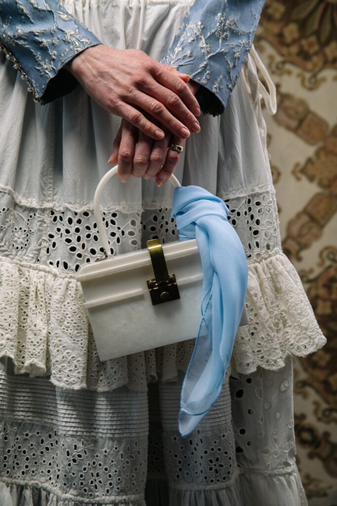 A woman holds a white bag with a blue scarf tied around it as she wears a spring white skirt.