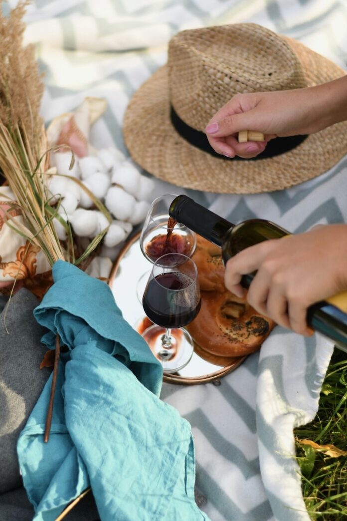 A person pours red wine into two glasses sitting on a picnic blanket beside a summer hat and sprigs of cotton.