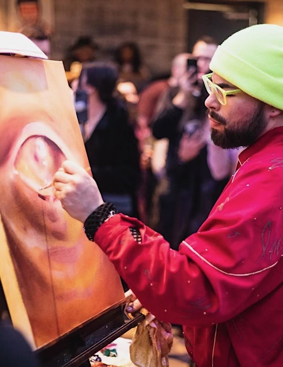 A man in a green beanie and red jacket paints an eye on a canvas at an art battle event in Pittsburgh.