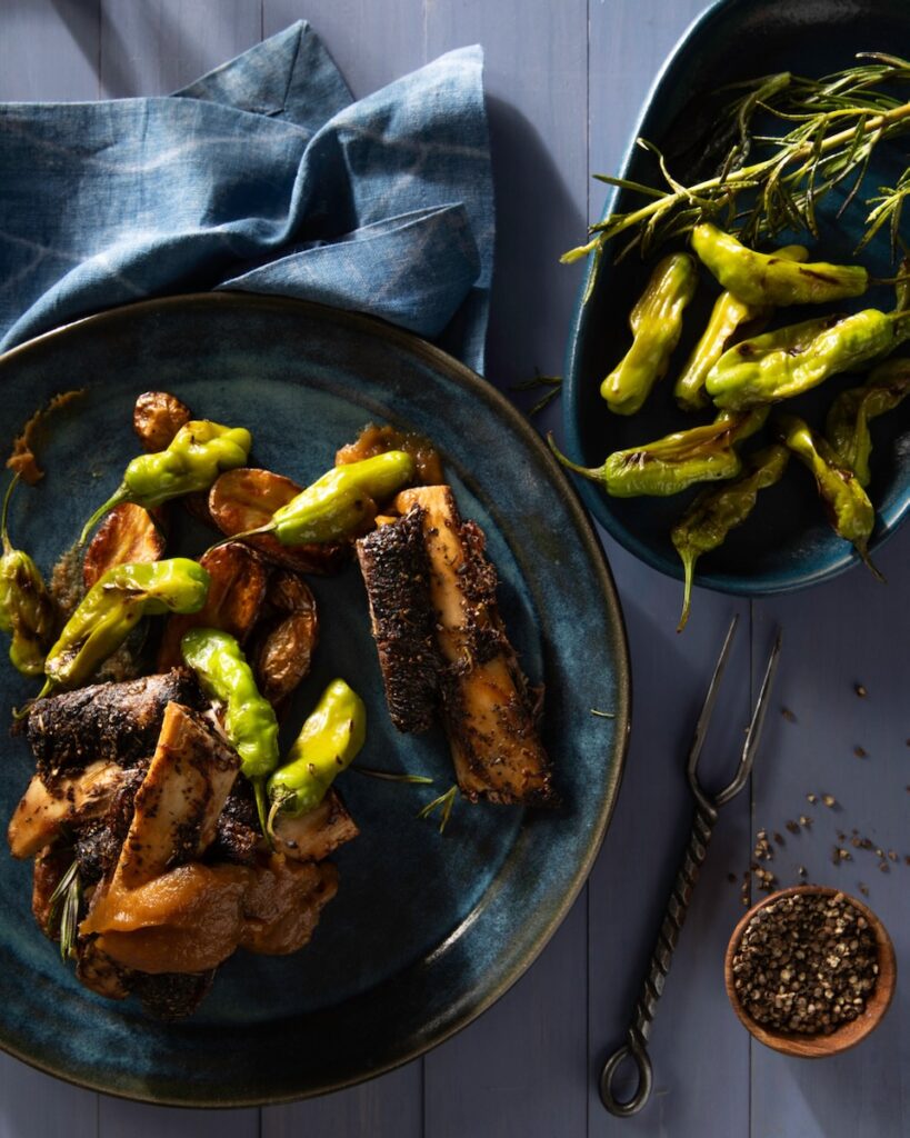 A blue plate with BBQ beef ribs covered in shishito peppers as a blue linen sits above the plate and a bowl of shishito peppers sits to the right of the plate.