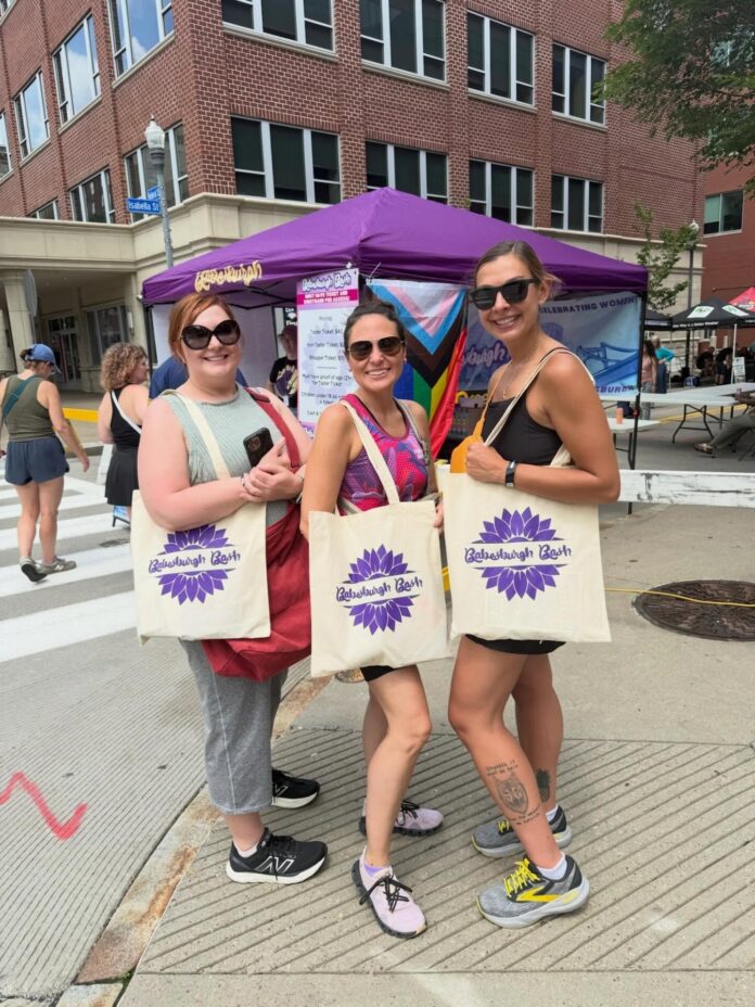 Three women with Babesburgh Bash tote bags and sunglasses on outside a purple vendor tent.