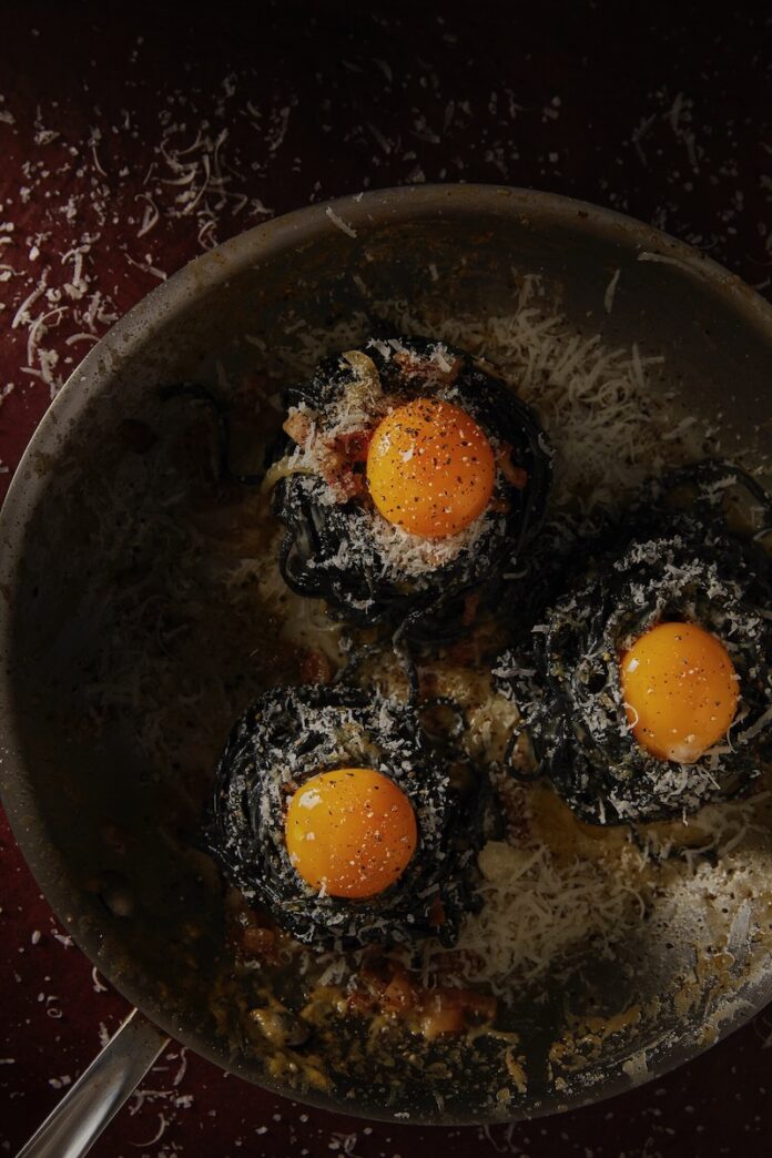 Carbonara with Handmade Charcoal Trenette Three bundles of handmade charcoal trenette made intoa. traditional Carbonara with yellow cheese on top of each pile.