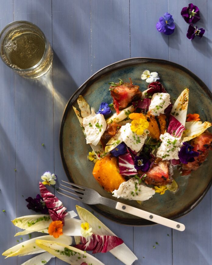 A green olive plate holds a charred heirloom tomato and mozzarella salad with various bright vegetables and a glass of sparkling water nearby.