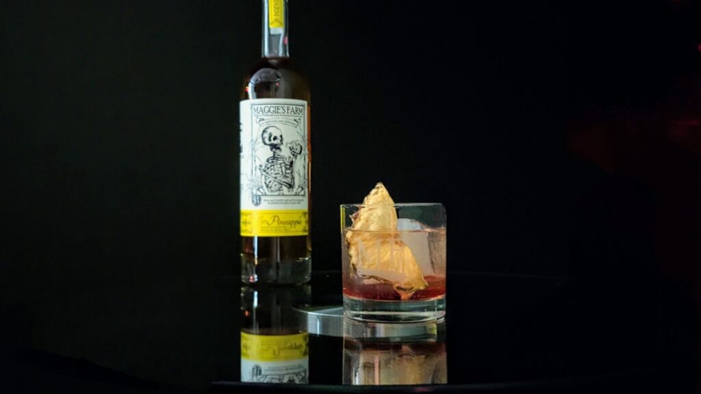 A bottle of Pittsburgh's Maggie's Farm Rum and a short cocktail glass sit beside each other on a black reflective table.