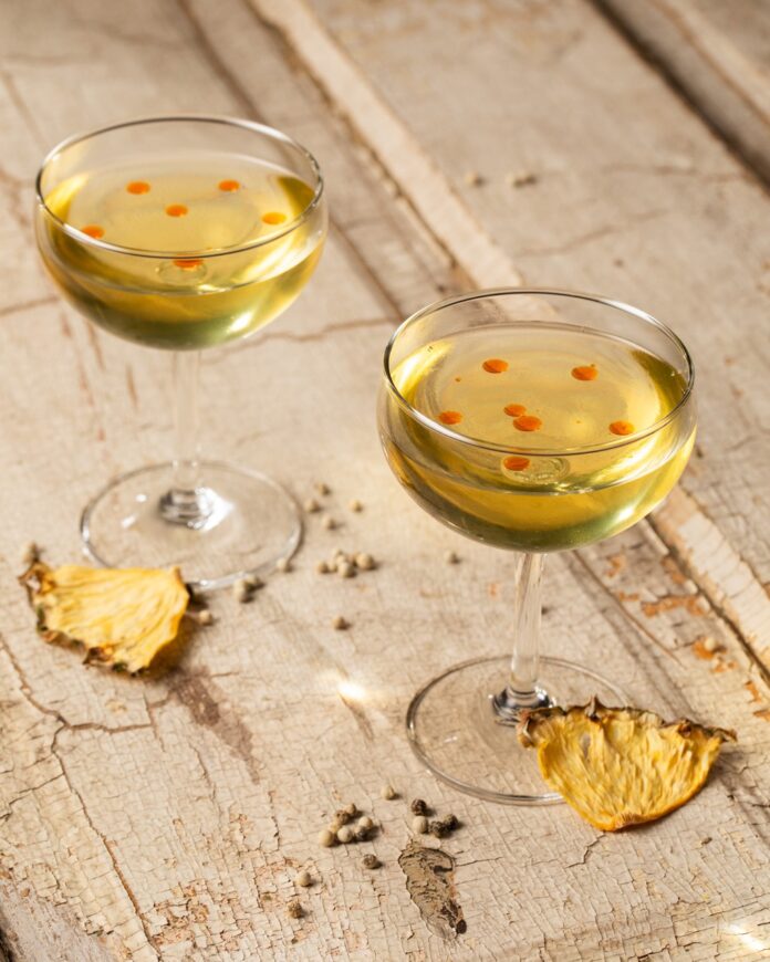 Two golden cocktails from a Pittsburgh bar with drops of orange oil on the inside and dried pineapple slices beside it.