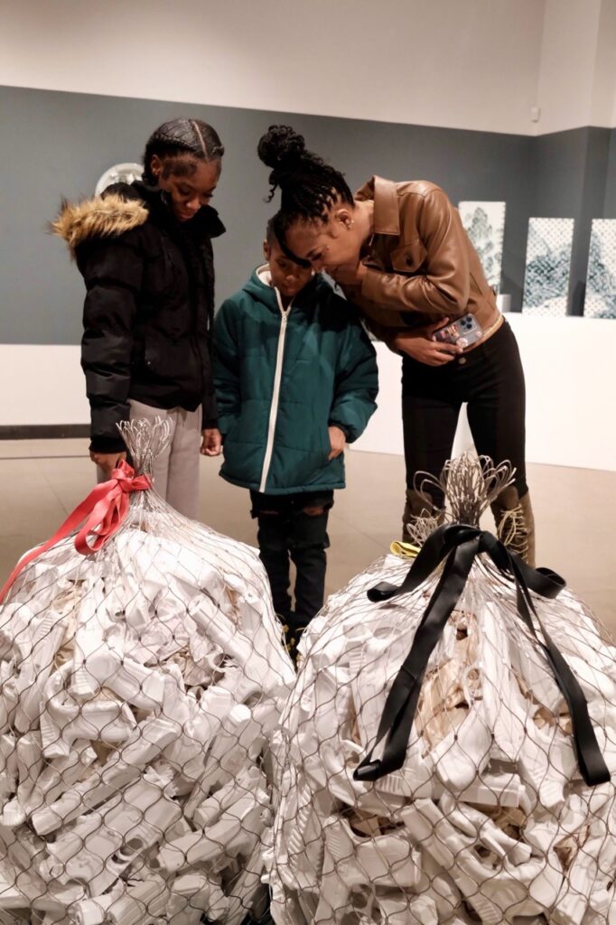 A woman and two younger girls look at a sculpture of chicken wire garbage bags.