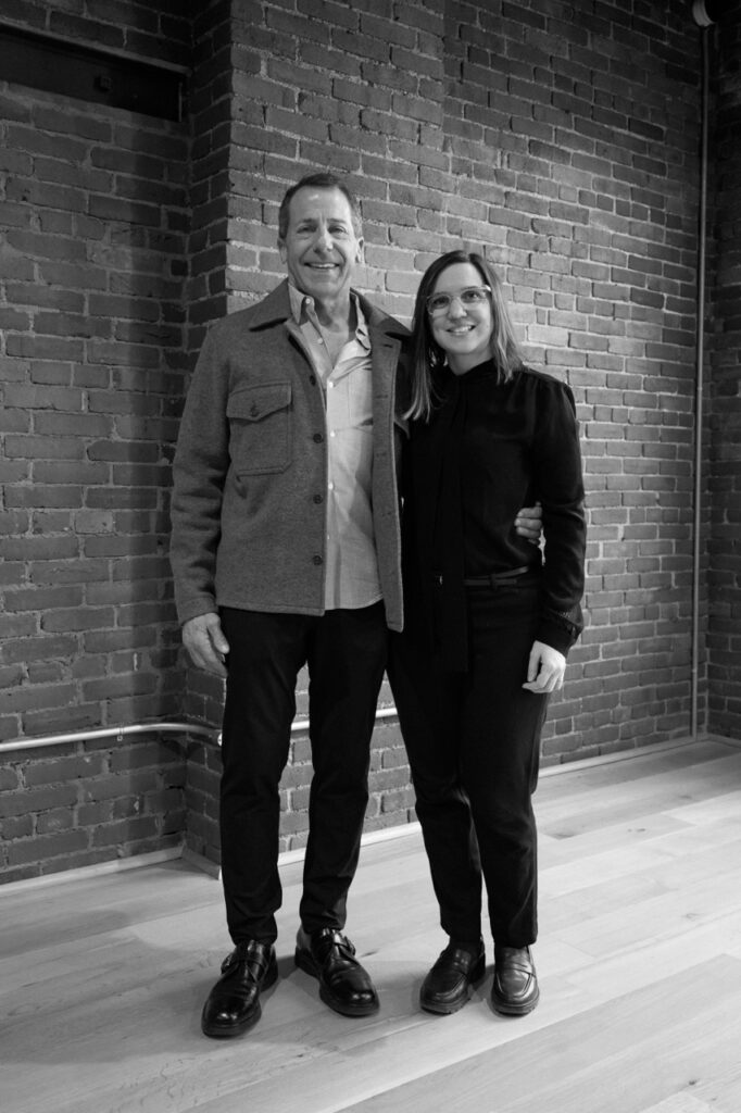 Two people stand arm in arm in front of a brick wall.