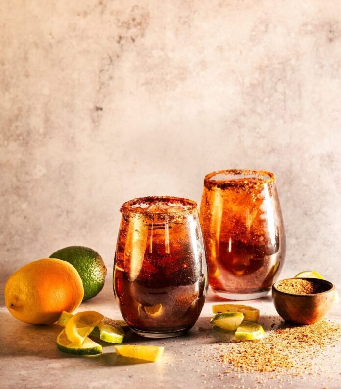 Dark and Handsome Rum Moscow Mule Two Dark and Handsome Rum Moscow Mules sit in front of a painted brown background with Tajin rims and a lime and orange sitting next to the glasses.
