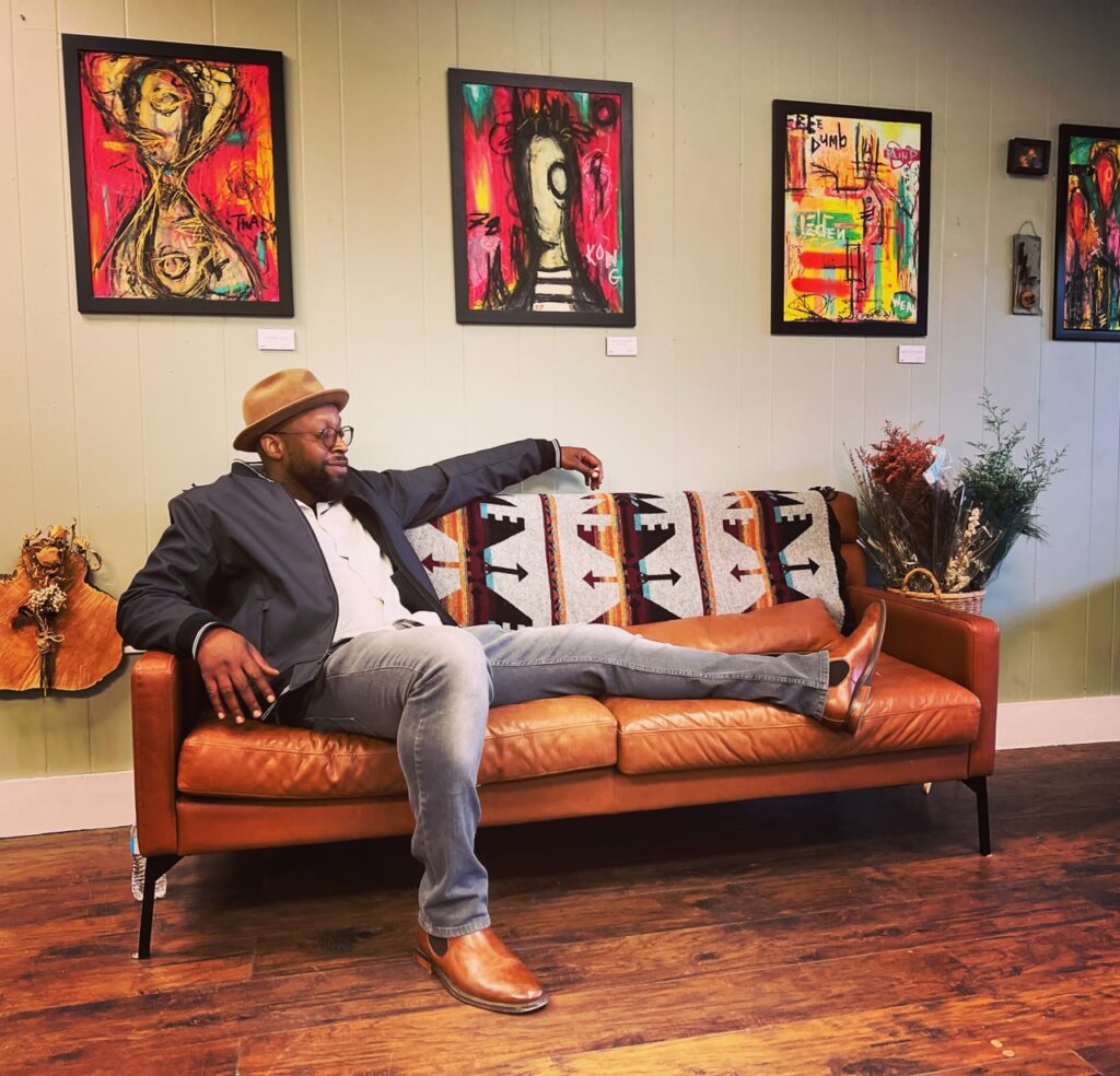 An emerging artist in Pittsburgh lounges on an orange sofa in a suit jacket and brown straw fedora.