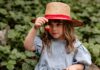 Your Strawberry Guide: From Foraging to Cooking A young girl in a blue dress and red sunhat sits on a bench holding a garden strawberry in front of her face with a basket of strawberries on her lap.
