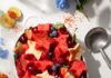 Plan Your Memorial Day Picnic A bow sits on a picnic table filled with star shaped watermelon, strawberries, blueberries, and spinkled with tajin to make a red white and blue fruit salad.