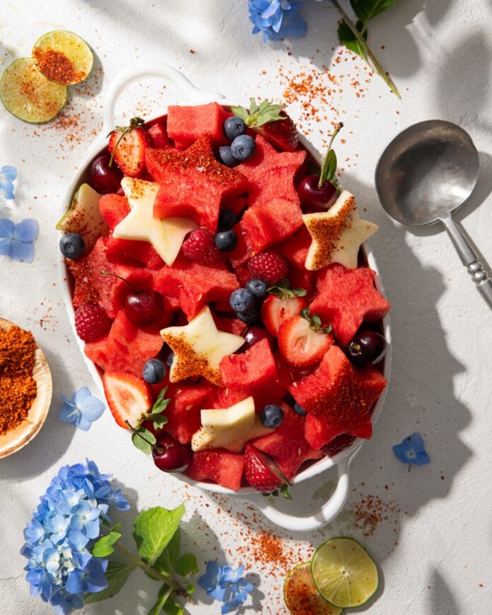 A bow sits on a picnic table filled with star shaped watermelon, strawberries, blueberries, and spinkled with tajin to make a red white and blue fruit salad.