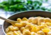 Go-To Cheddar Mac and Cheese A blue pot of cheddar shell mac and cheese in a yellow color with a wooden spoon stuck inside it.