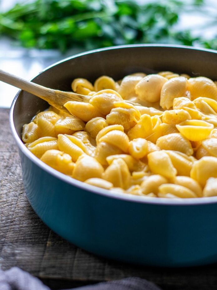 Go-To Cheddar Mac and Cheese TABLE Magazine A blue pot of cheddar shell mac and cheese in a yellow color with a wooden spoon stuck inside it.