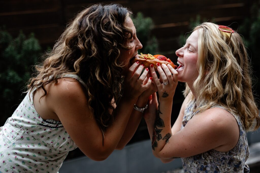 Two women share one hot dog at a side profile.
