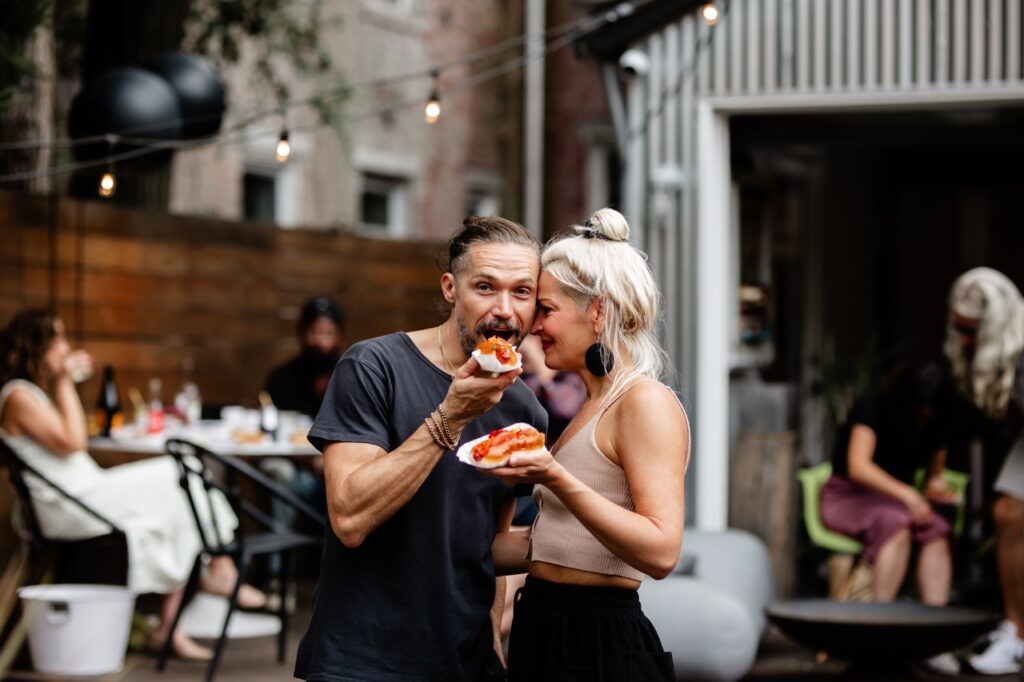 A man bites into a hot dog while a woman holding a hot dog leans her face against his.
