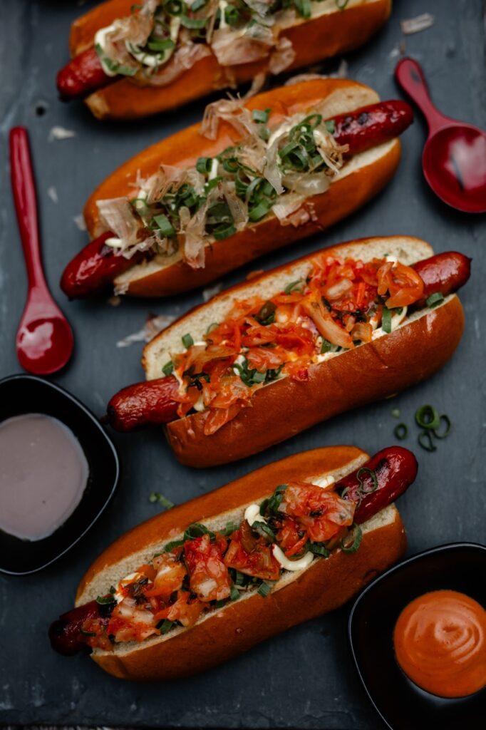 Four hot dogs with various gourmet toppings sit on a blue table beside two spoons.