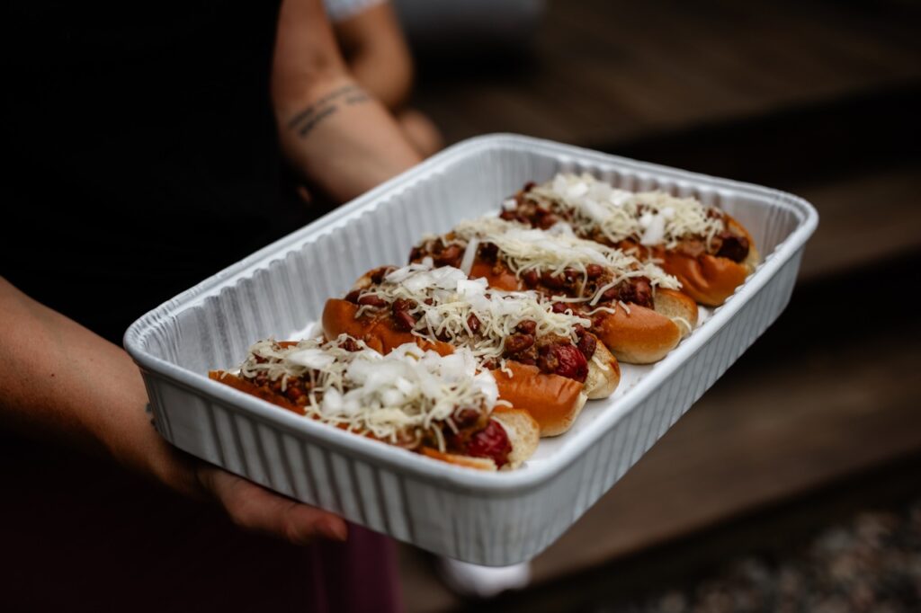 A person carries a pan full of four gourmet hot dogs.