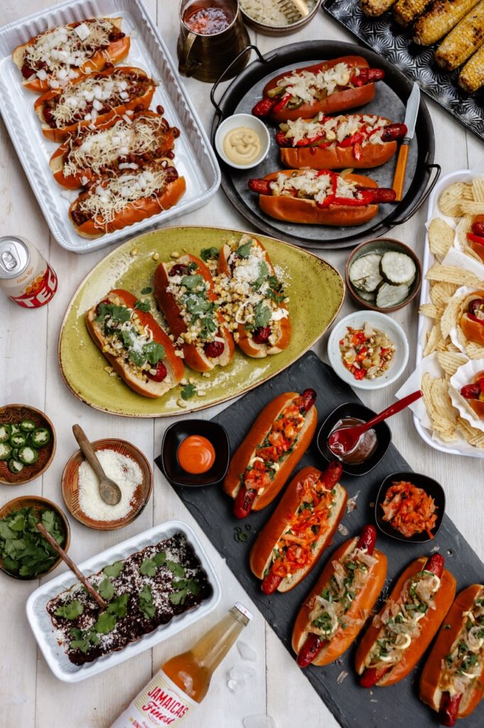Various plates of gourmet hot dogs with various vegetable and condiment toppings sit around the containers of toppings and sauces.