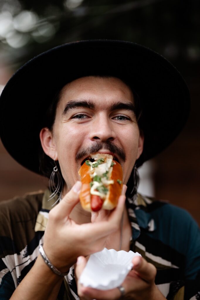 A man in a black hat bites into a gourmet hot dog.