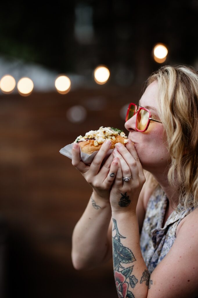 A side profile of a woman with red glasses bites into a gourmet hot dog.