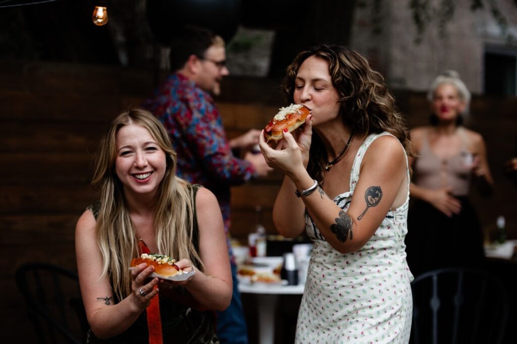 One woman smiles with a hot dog while another in white bites into her hot dog.