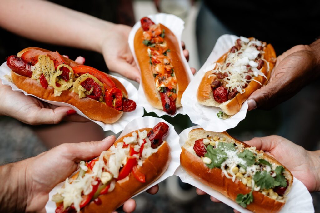 Five people hot gourmet hot dogs with various green, white, and red toppings.