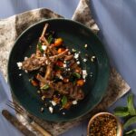 A green plate holds a grilled lamb legs scottadito with golden fork and knife beneath the plate and a small container of peppercorns on the other side.