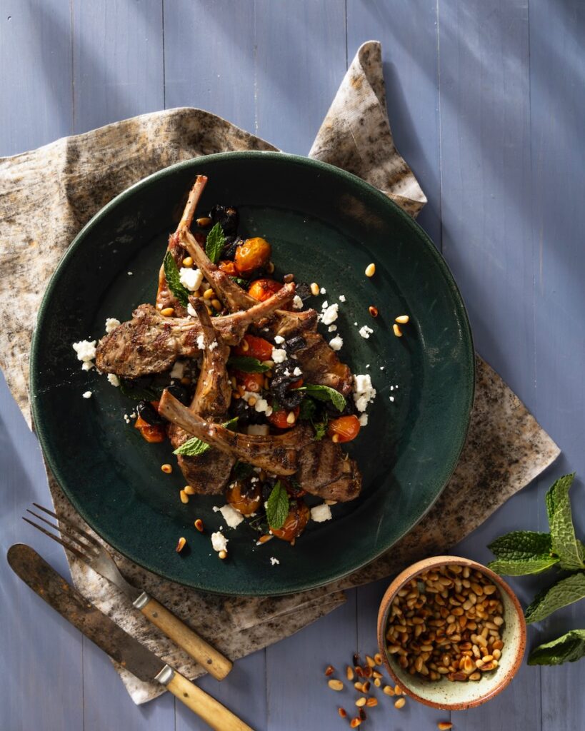A green plate holds a grilled lamb legs scottadito with golden fork and knife beneath the plate and a small container of peppercorns on the other side.
