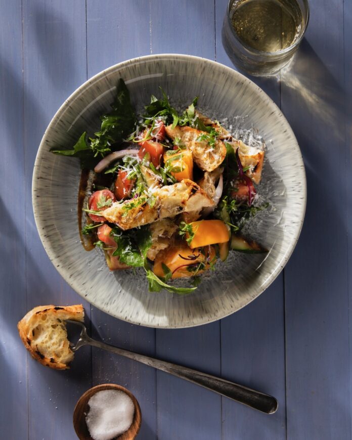 A white bowl full of a grilled Panzanella Salad with onions, tomatoes, and zucchini inside, all sitting on a blue picnic table with a spoon sitting below the bowl.