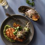 On a green plate sits a grilled salmon steak on a bed of salsa verde with a glass of wine to the top left and side tray that matches the plate to the right.