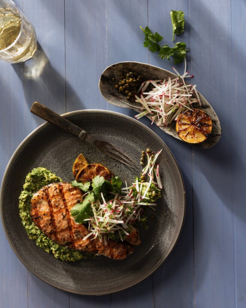 On a green plate sits a grilled salmon steak on a bed of salsa verde with a glass of wine to the top left and side tray that matches the plate to the right.