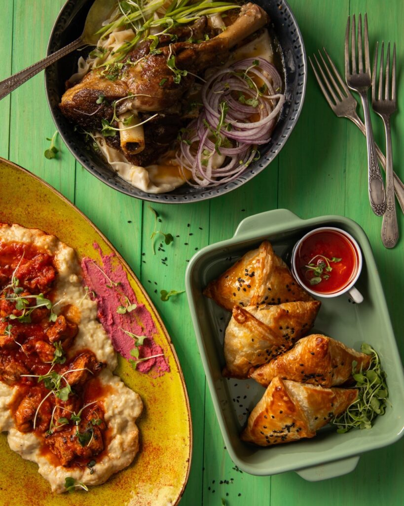 On top of a green picnic table sits three international restaurant dishes like rice and beef, stew, and deep fried dough pockets.