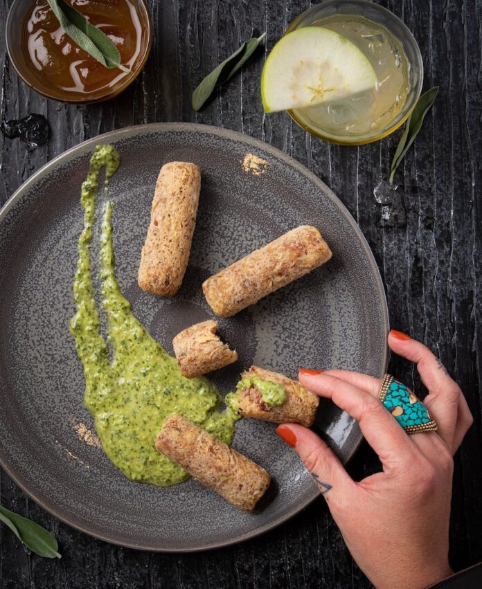 Lamb Croquettes A woman with a ring on her finger picks up a log of Lamb Croquettes off of a grey plate with a green sauce smeared across it.