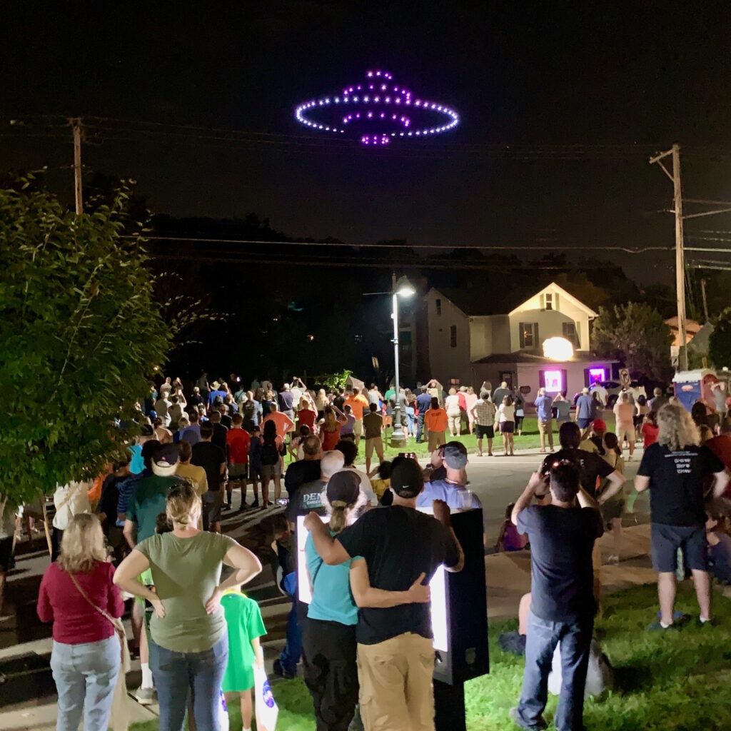 A group of people on a Pittsburgh lawn look up at a UFO shape in the sky made out of drones.