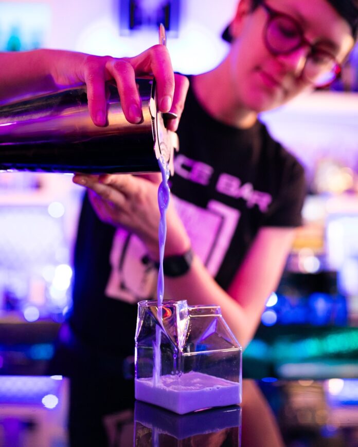 A bartender pours a mocktail from a cocktail shaker into a clear milk jug glass.