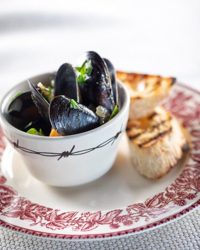 Mussels in Broth A small bowl hols Mussels in a broth that sit on a plate with two pieces of toasted baguette beside the bowl.