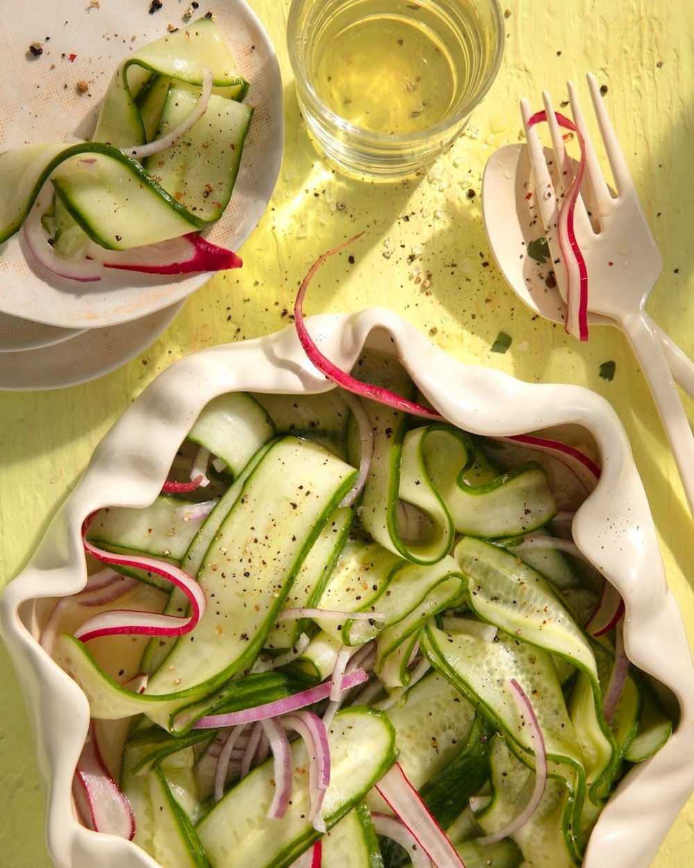 a bowl filled with Nana’s Cucumber Salad next to a smaller bowl and a fork and a knife