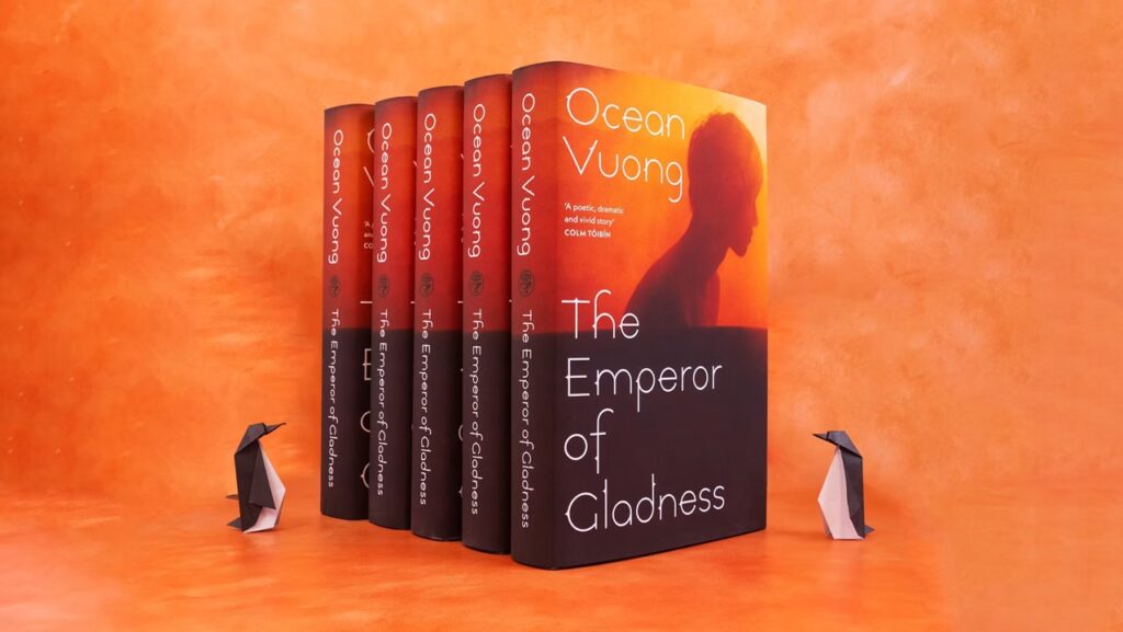 An orange background with 5 books from an author visit Pittsburgh in front of it and two paper made penguins on each side.