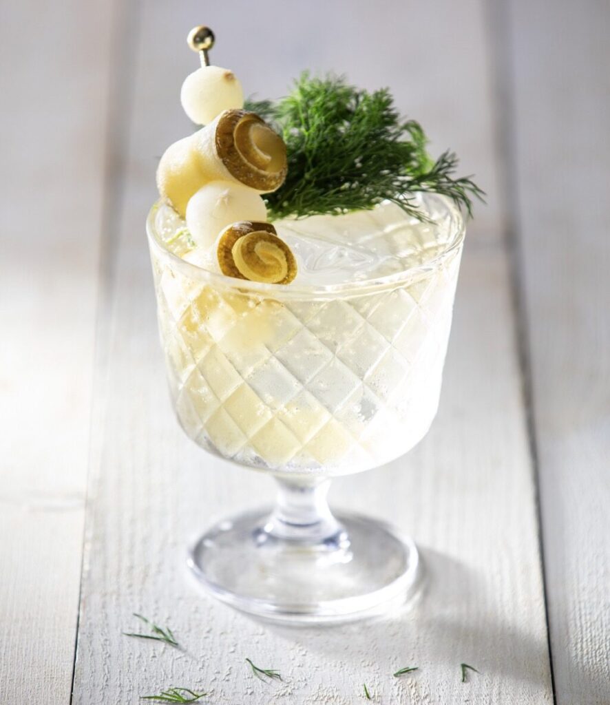 A short glass holds a pickle spritz on a white picnic table topped with rolled pickles, cocktail onions, and fresh dill.