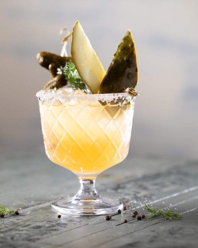 Lowball glass with a pickleback mocktail inside topped with pickle spears and kosher salt on the rim as it sits on a grey table.
