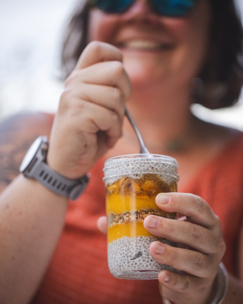 A woman dips a spoon into a chia seed and peach parfait with a smile on her face at a Pittsburgh park picnic.