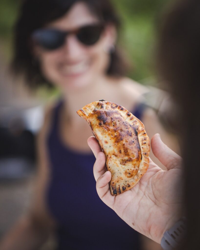 A woman in a purple tank top smiles in the background as a woman holds an empanada up to the camera.