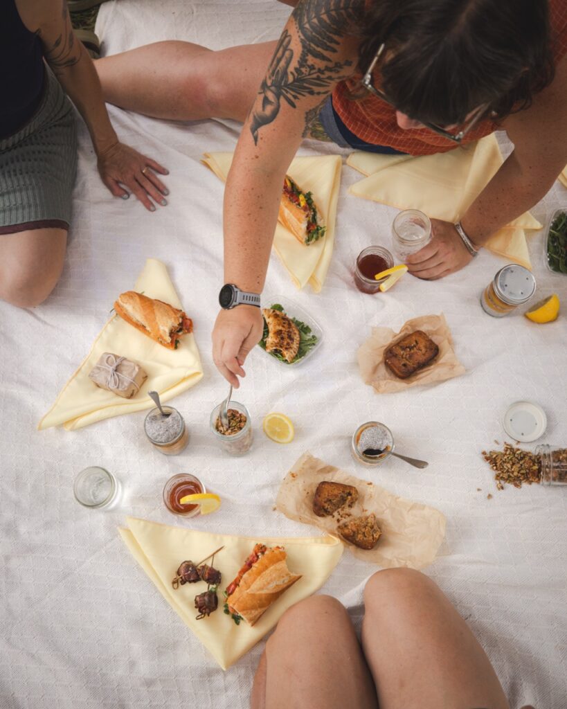 Three spot sit around a white picnic blanket digging into various foods.