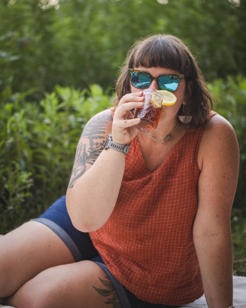 A woman in an orange tank top and sun glasses sips iced tea at a picnic in a Pittsburgh park.