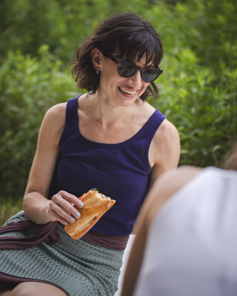A woman in a purple tank top and sunglasses smiles and eats a sandwich at a picnic in a Pittsburgh park.
