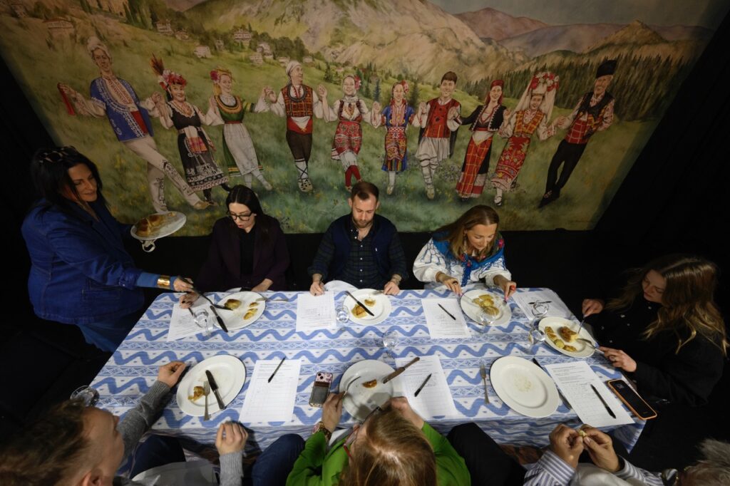 Seven people sit at a blue and white table as a woman in blue serves them pierogis.