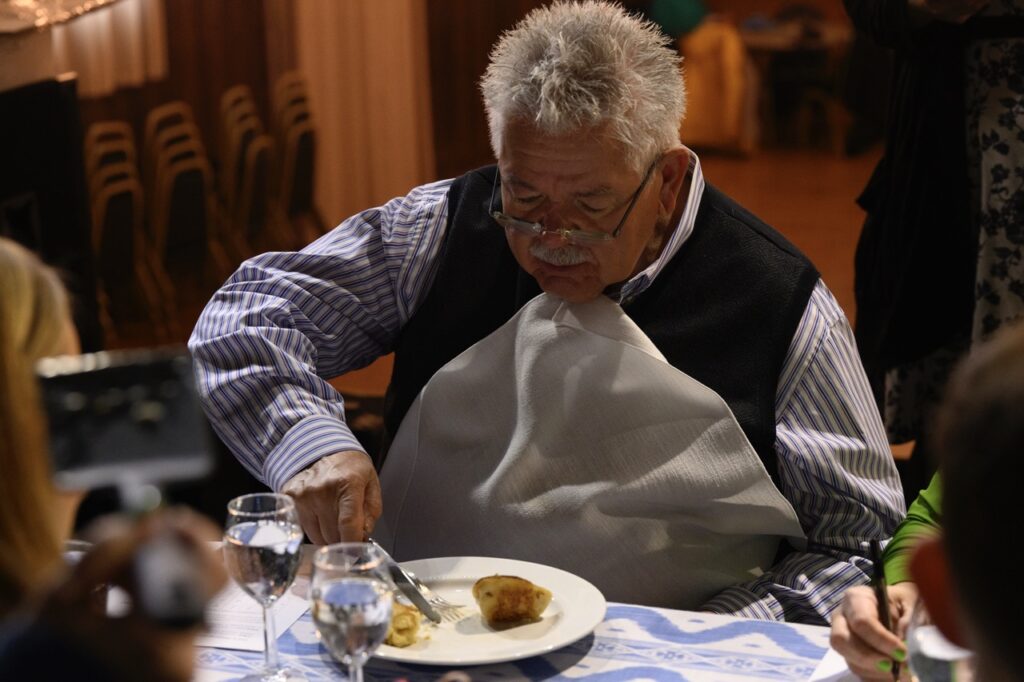 A man in a long sleeve shirt and vest cuts up a pierogi on his plate.