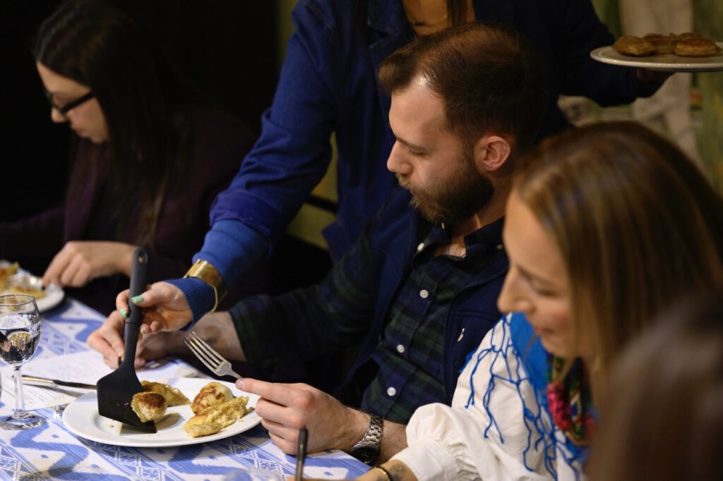 A person serves a man a pierogi from a kitchen spoon.